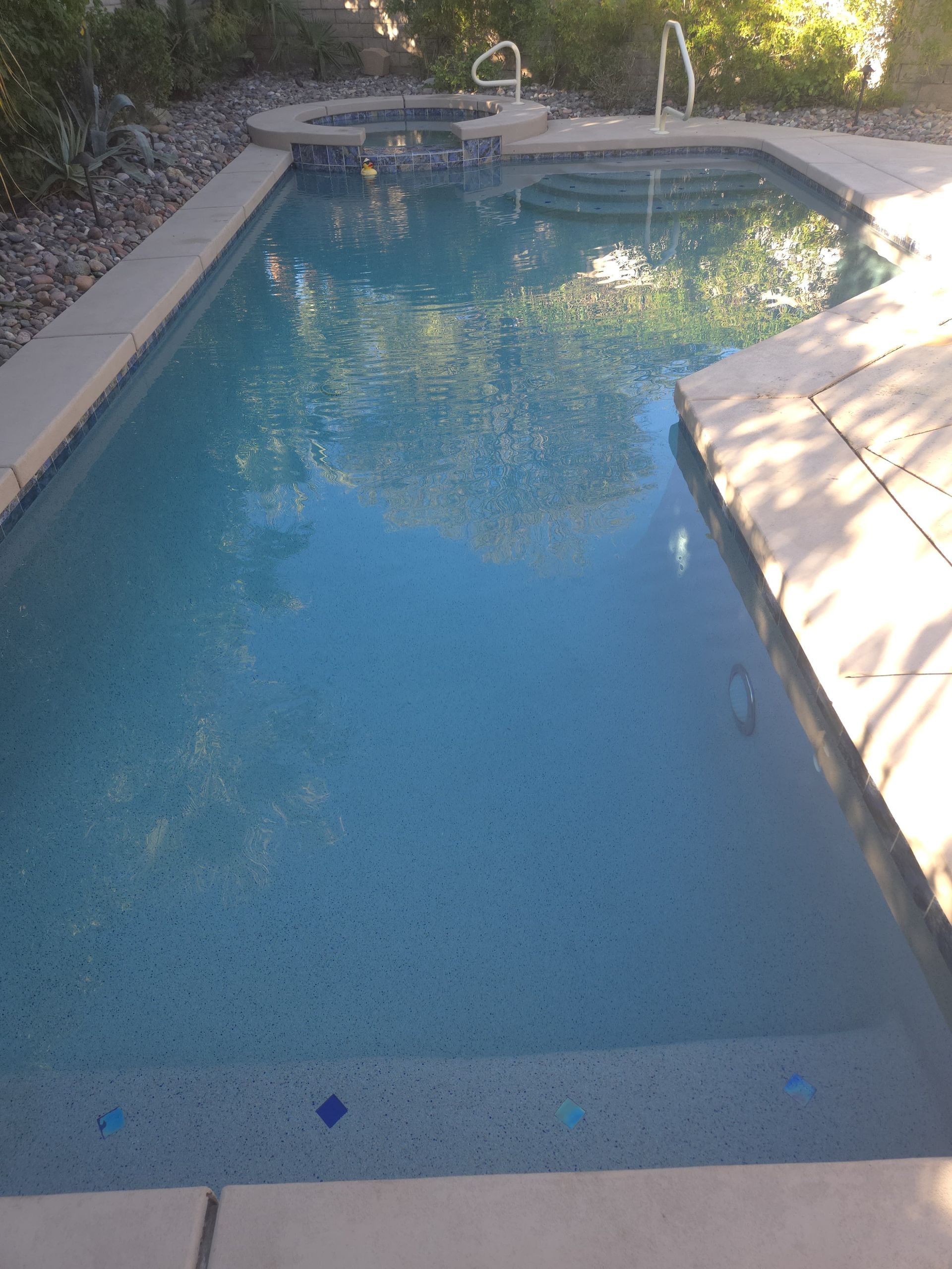 A rectangular swimming pool with a connected jacuzzi. Blue water, light concrete edges.