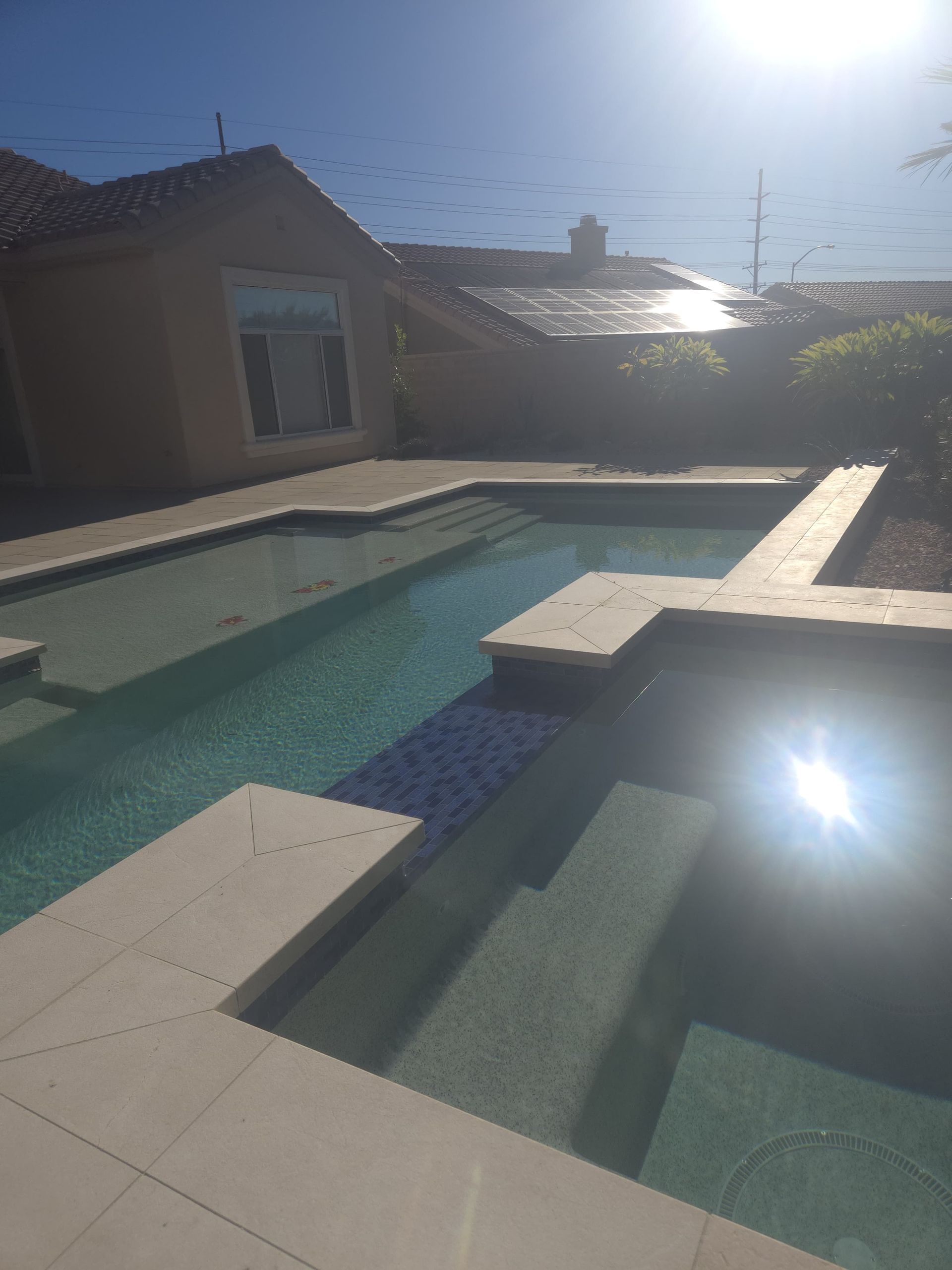 Pool area with light-colored tile and steps. Sunlight reflects on the water. House with solar panels in the background.