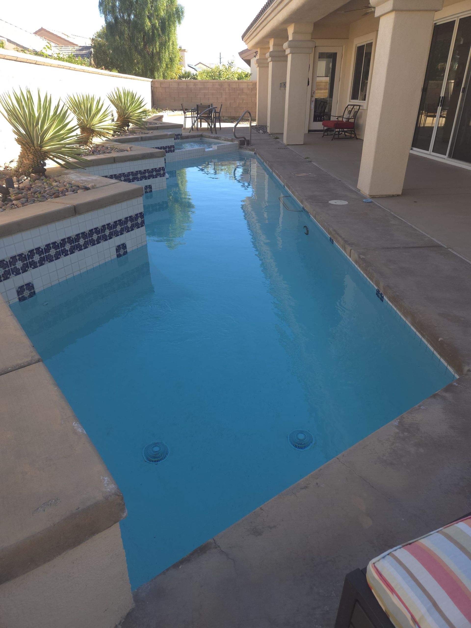Pool with blue water next to a covered patio. White columns, stone deck, blue and white tile.