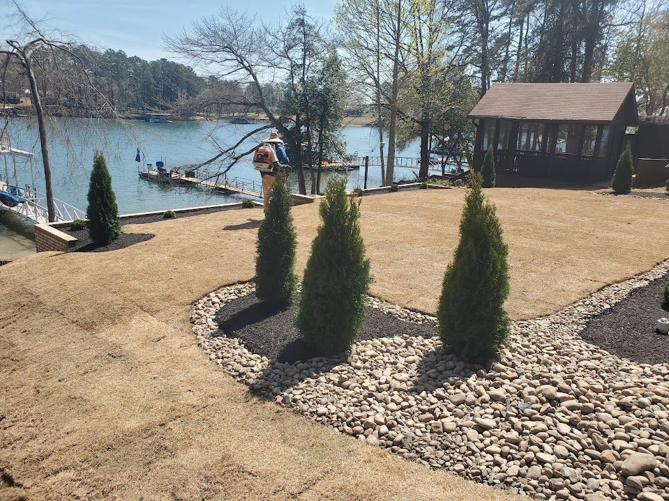 a lawn with trees and newly installed river rocks next to a body of water