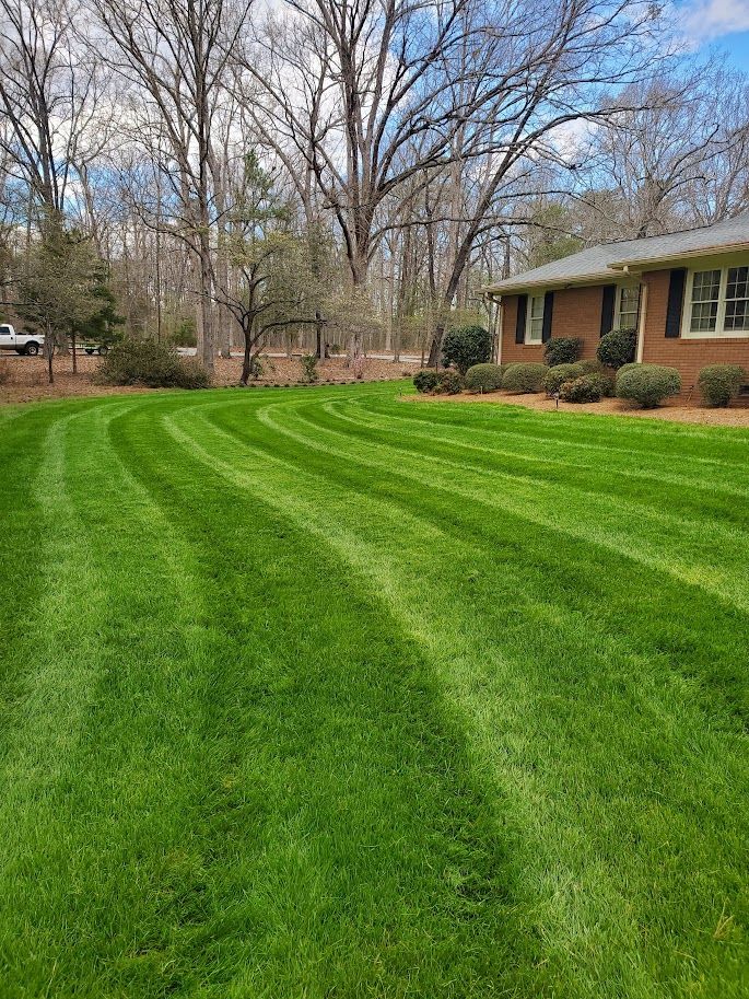 a lush and freshly mowed green lawn in front of a brick house with trees in the background 