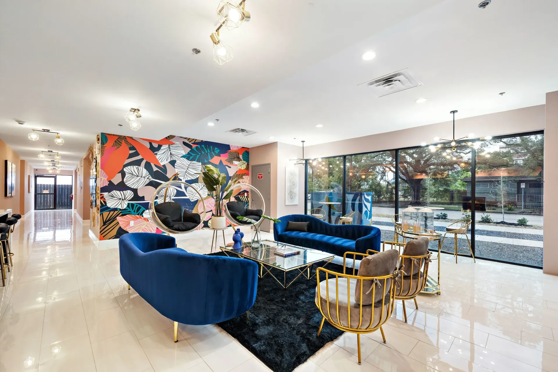A modern, colorful living space with blue couches, gold chairs, and a mural. Large windows overlook a natural landscape.