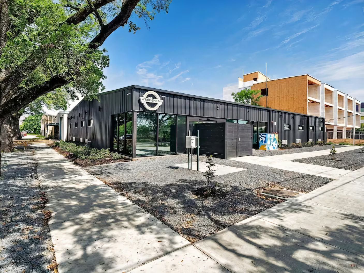 Edificio moderno de color negro con grandes ventanales y patio de grava bajo un cielo azul brillante.
