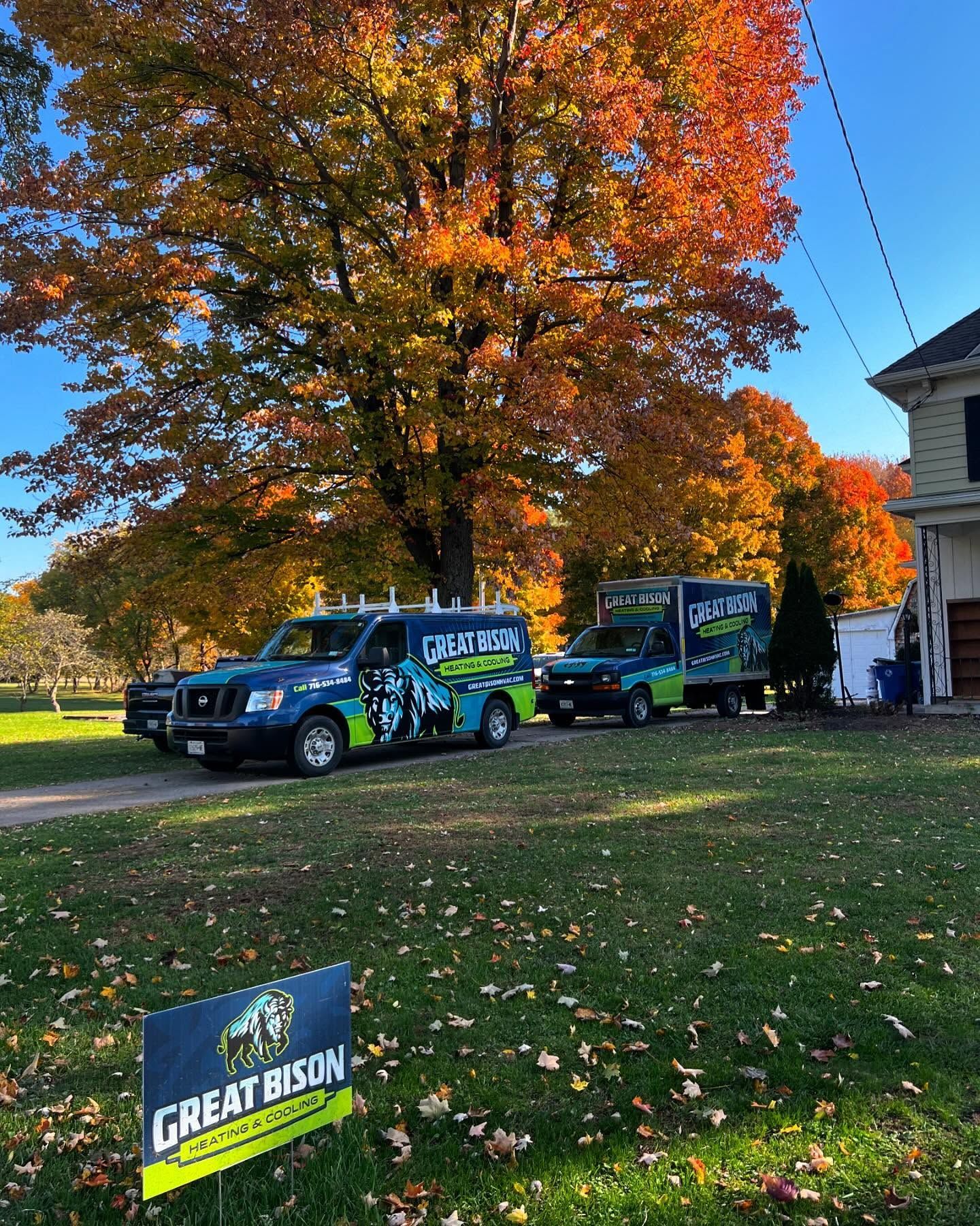 Two service trucks parked on a lawn with a sign. Fall foliage in the background.
