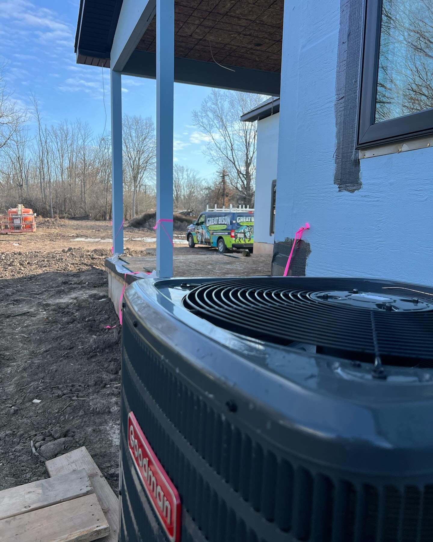 An air conditioning unit in front of a house under construction. A service van is parked in the distance.