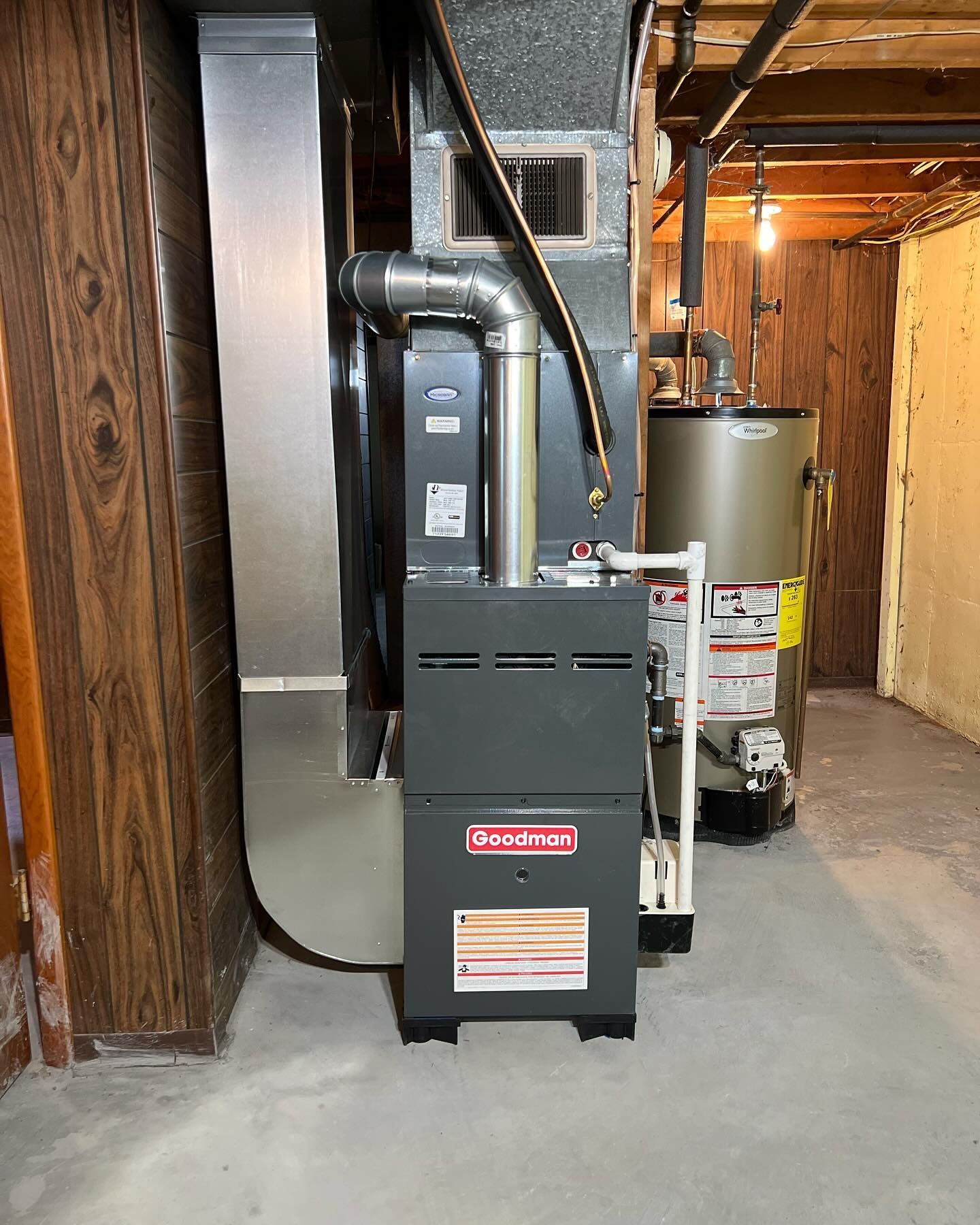 Furnace and water heater in a basement. Gray Goodman furnace, silver ductwork, and a brown water heater.