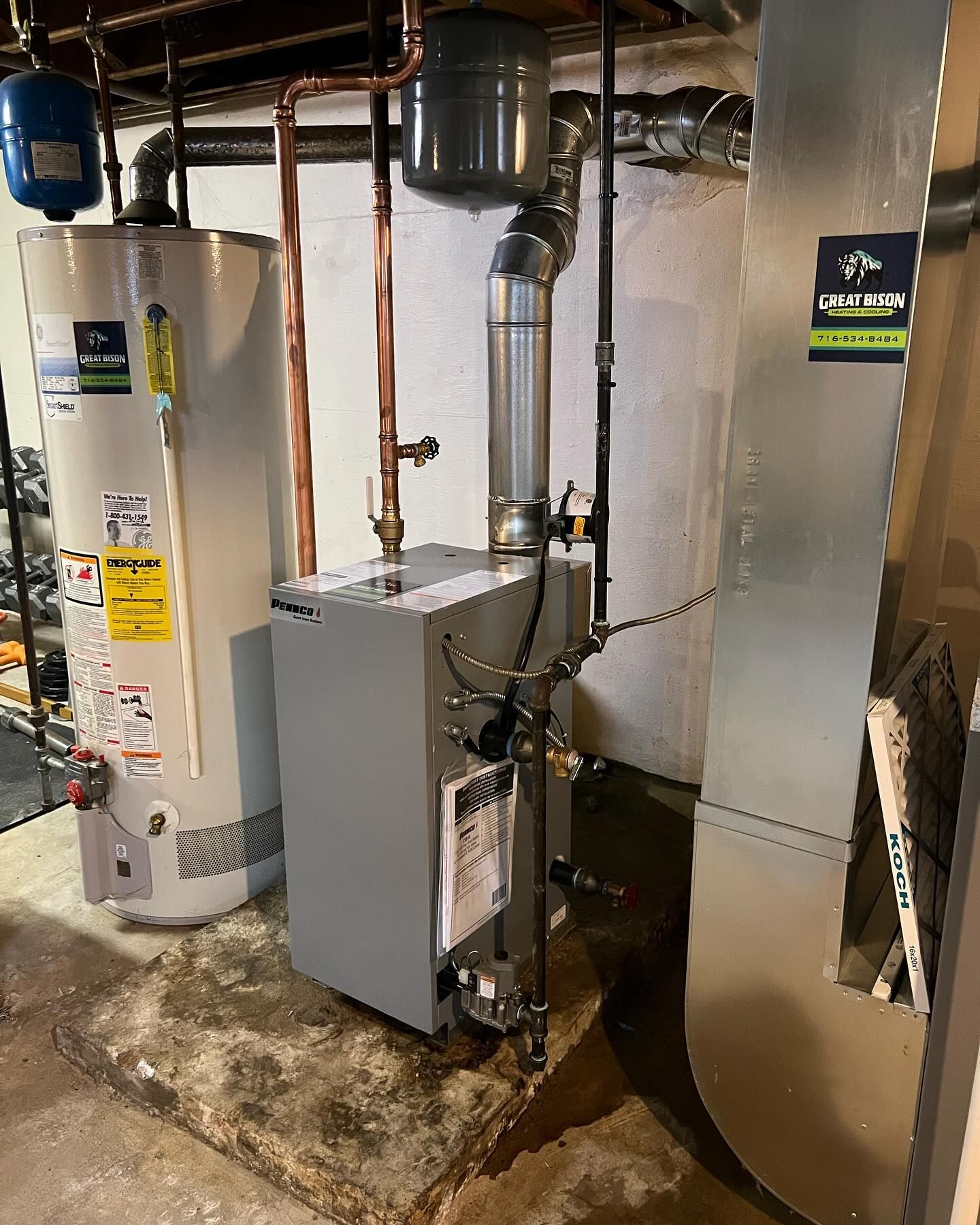 Water heater and furnace in a basement, connected by pipes. Gray metal and white walls.