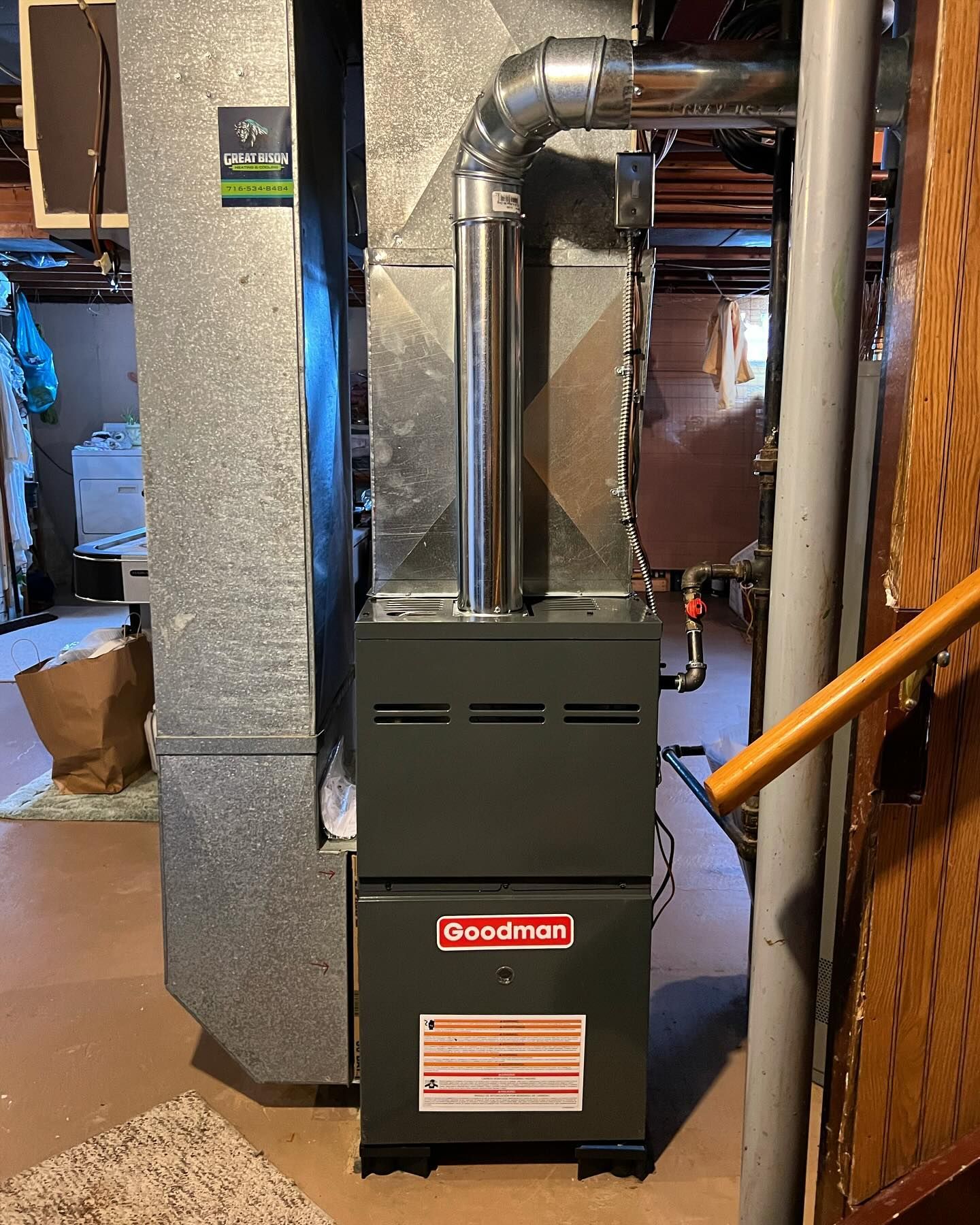 A Goodman furnace in a basement, with a silver exhaust pipe and ductwork, near a wooden door and stairs.