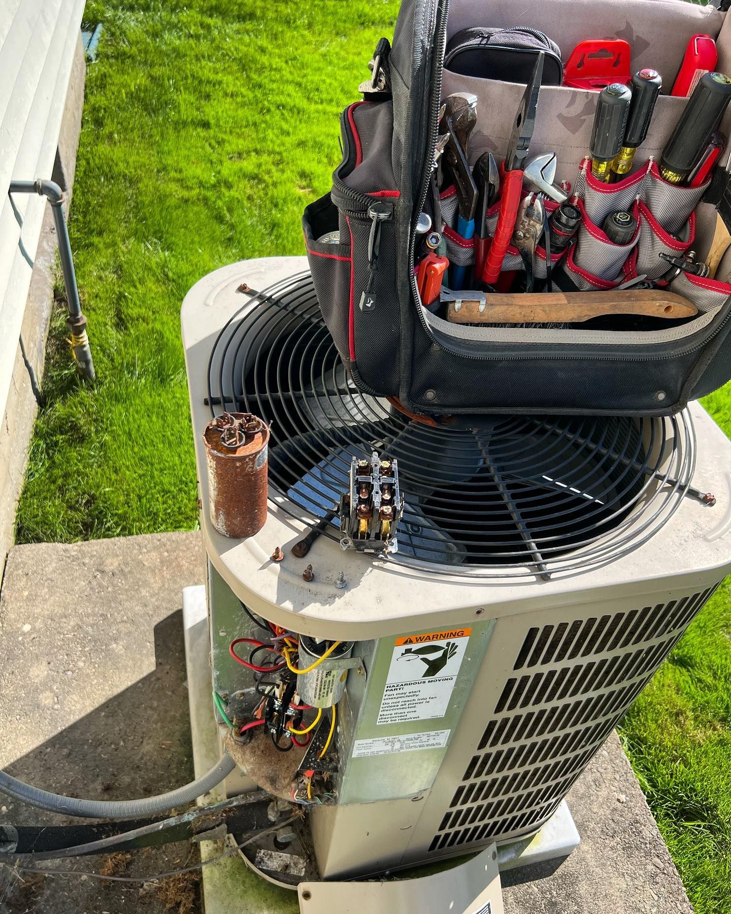 HVAC repair, tools in bag on top of an air conditioning unit. Green grass and building in background.