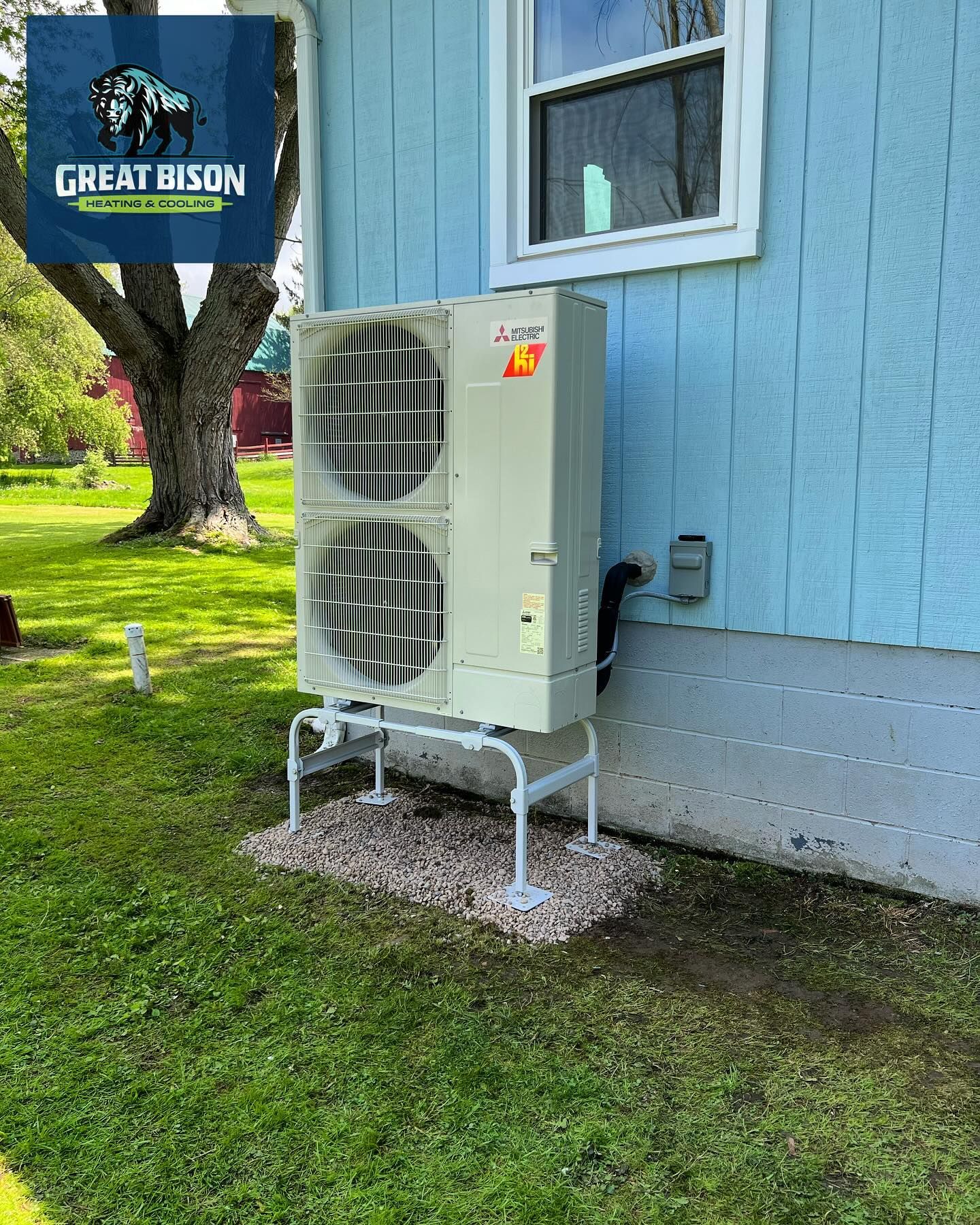 Outdoor heat pump unit mounted on a stand next to a blue house and a grassy yard.