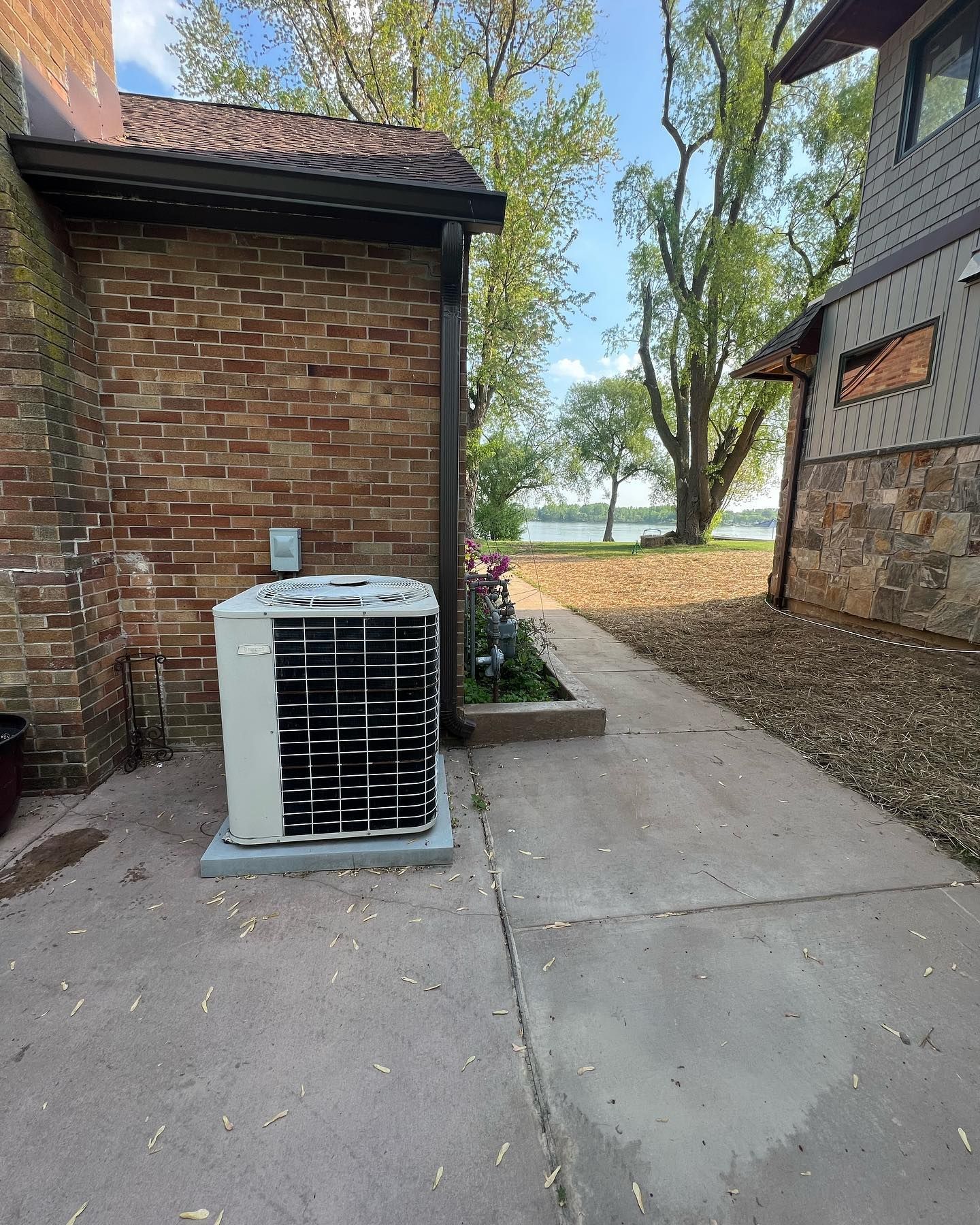 An air conditioning unit sits outside a brick building next to a concrete path leading to a lake.