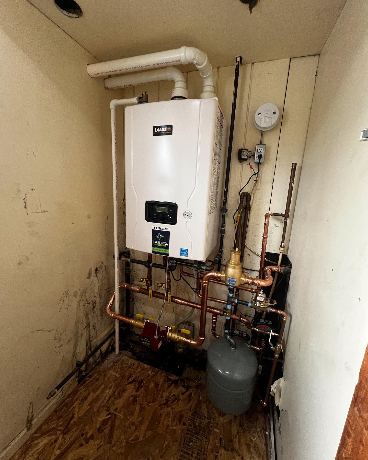 White wall-mounted furnace in a utility room with copper pipes, a gray expansion tank, and ventilation.