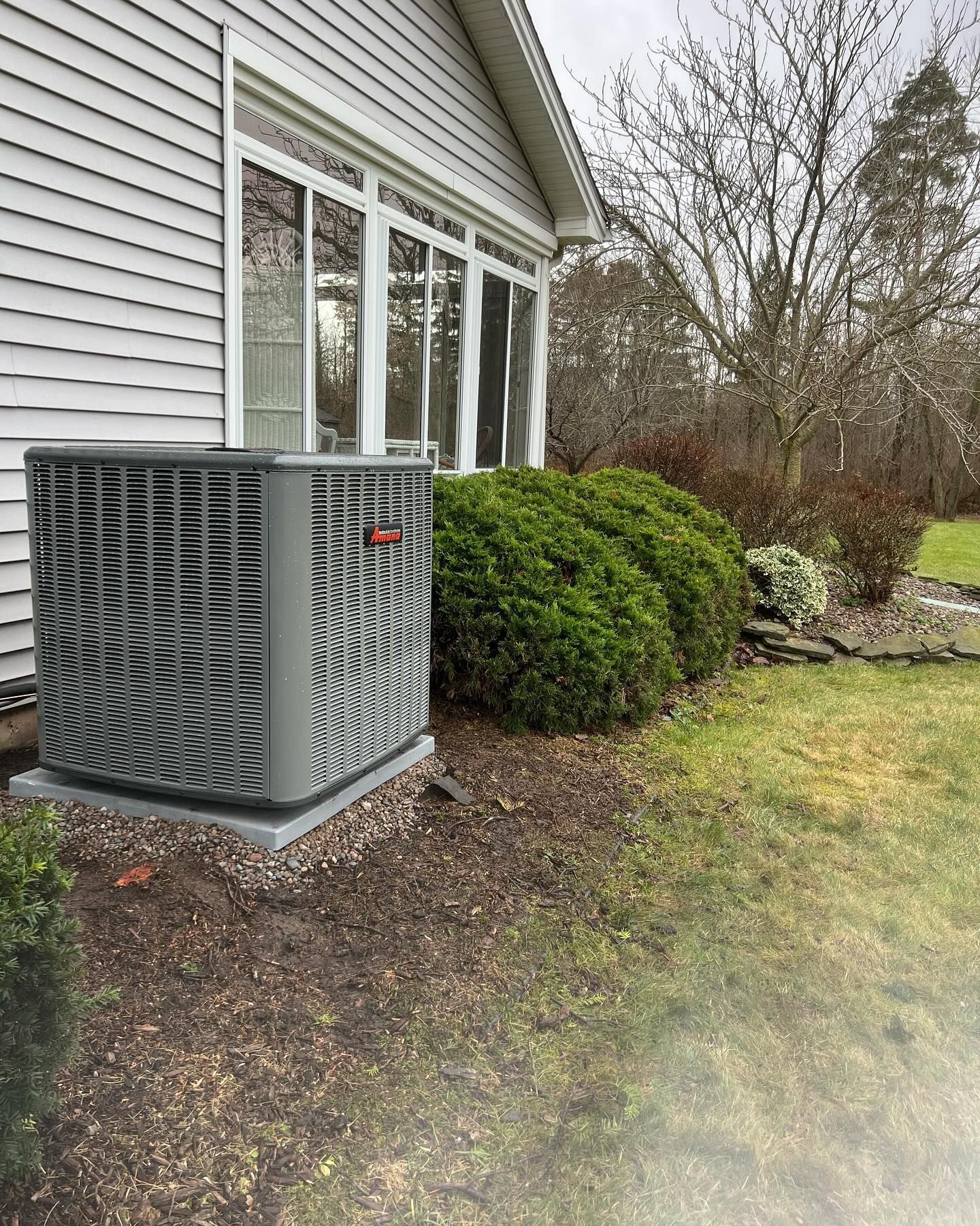 Gray AC unit by a house, next to green bushes. Cloudy day.
