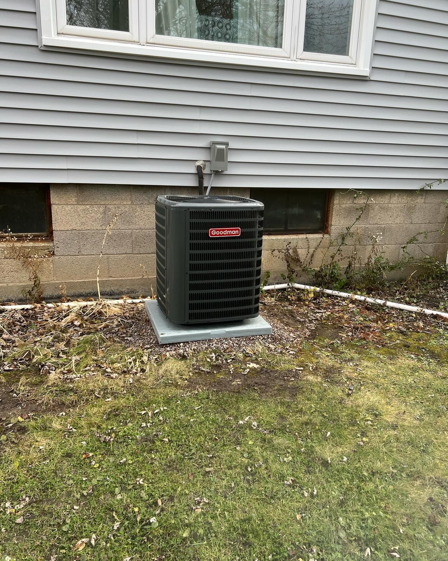 An outdoor Goodman air conditioning unit on a concrete pad near a house with gray siding.