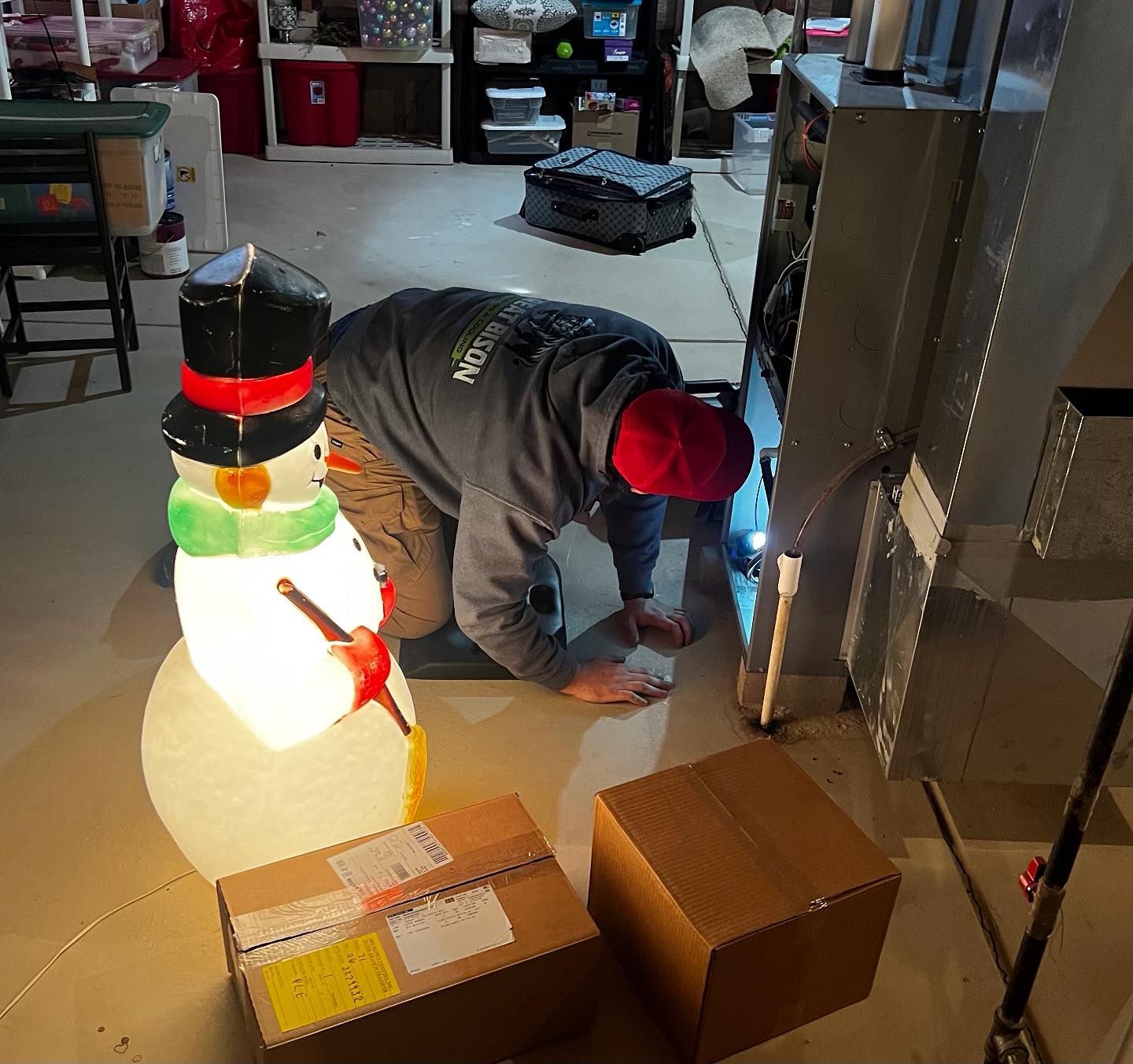 Man in a red hat working on something near a furnace in a basement, with a snowman decoration nearby.