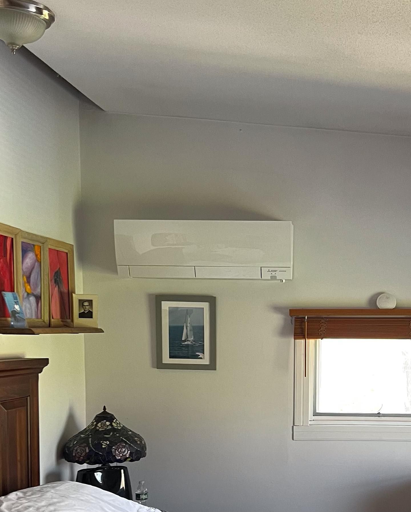 Wall-mounted air conditioner unit above a bed in a bedroom with art and a window.