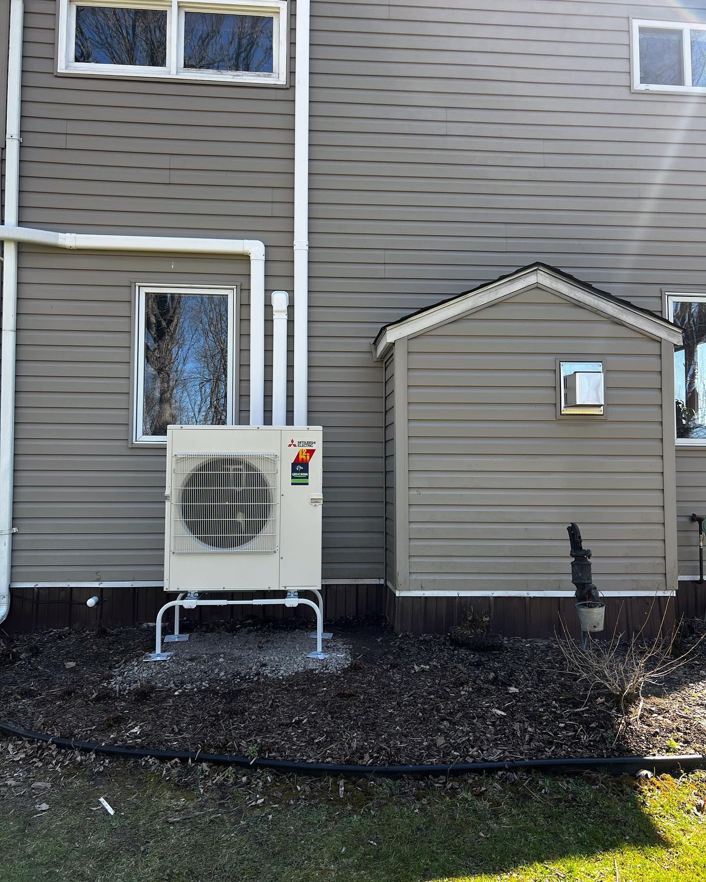 Heat pump unit next to a shed on the side of a house. Gray siding and small window visible.