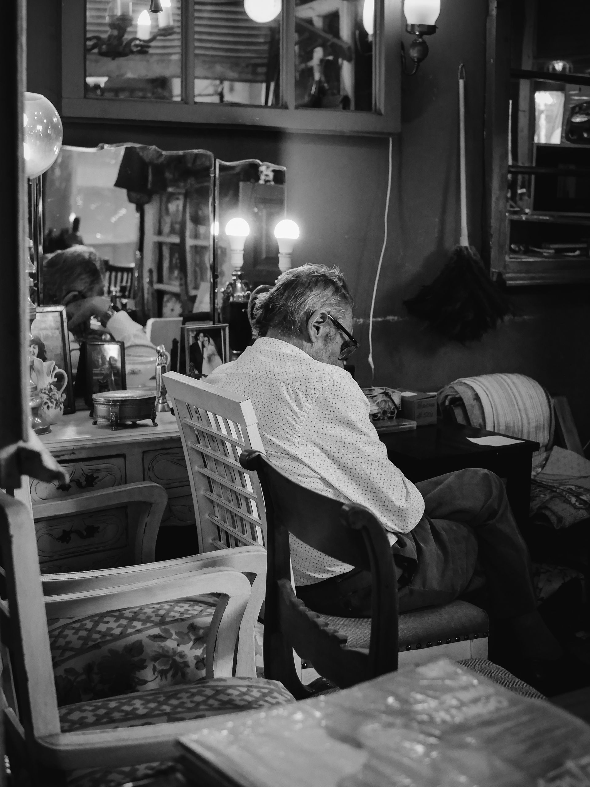 A person sitting in a chair in a dimly lit room, viewed from behind, with a mirror reflecting their back and head.