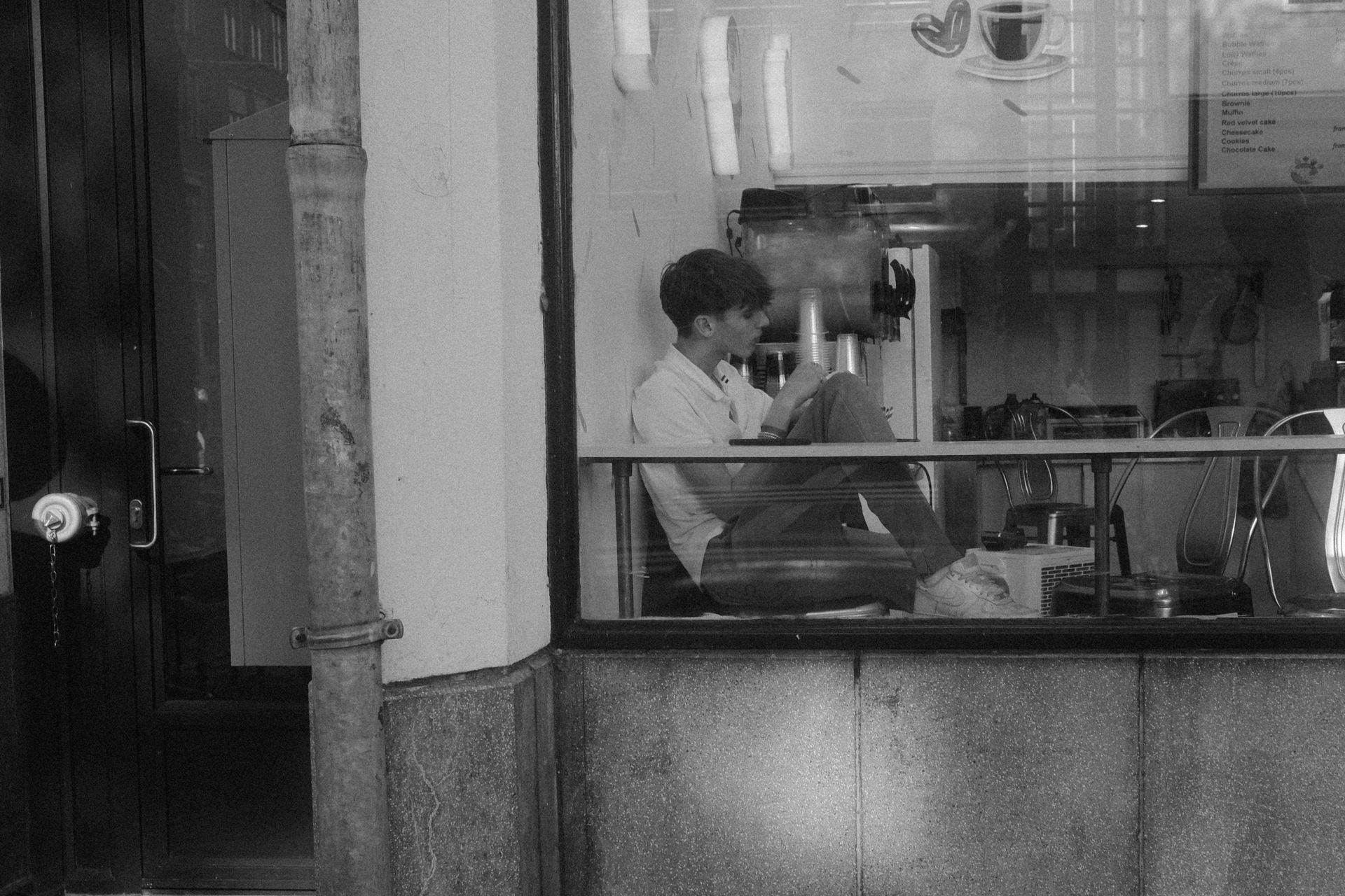 A person sits inside a cafe by a window, looking down at a book or device, captured in a monochrome street shot.