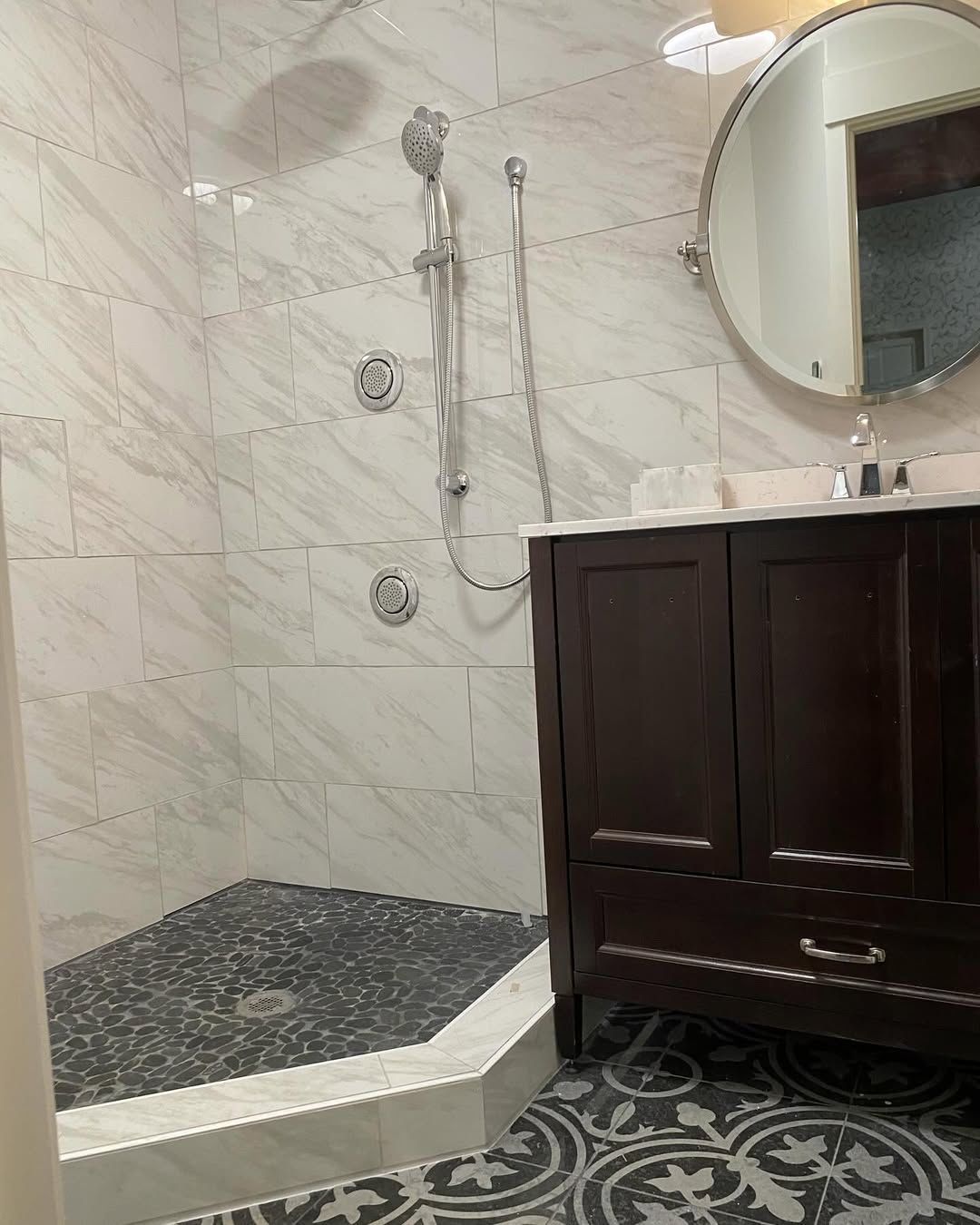 Bathroom with marble-tiled shower, dark vanity, and black stone shower floor. Round mirror and patterned floor tiles.