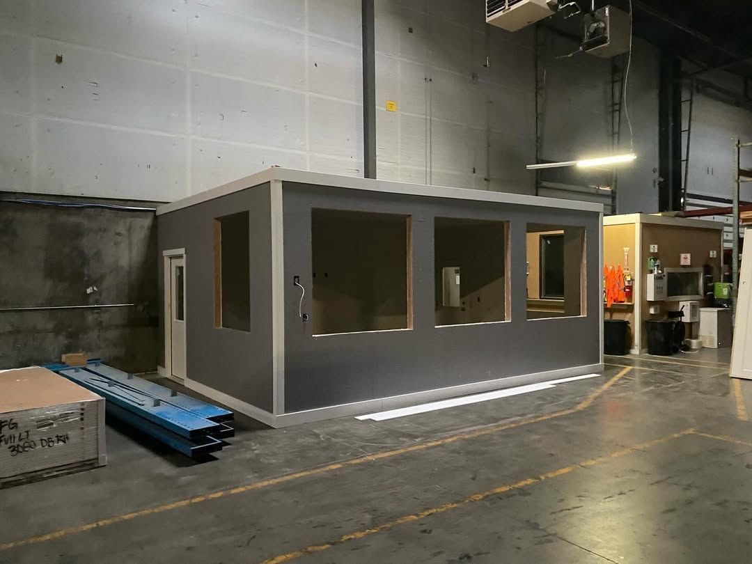 A small, gray building with large window openings inside a warehouse, with construction materials nearby.