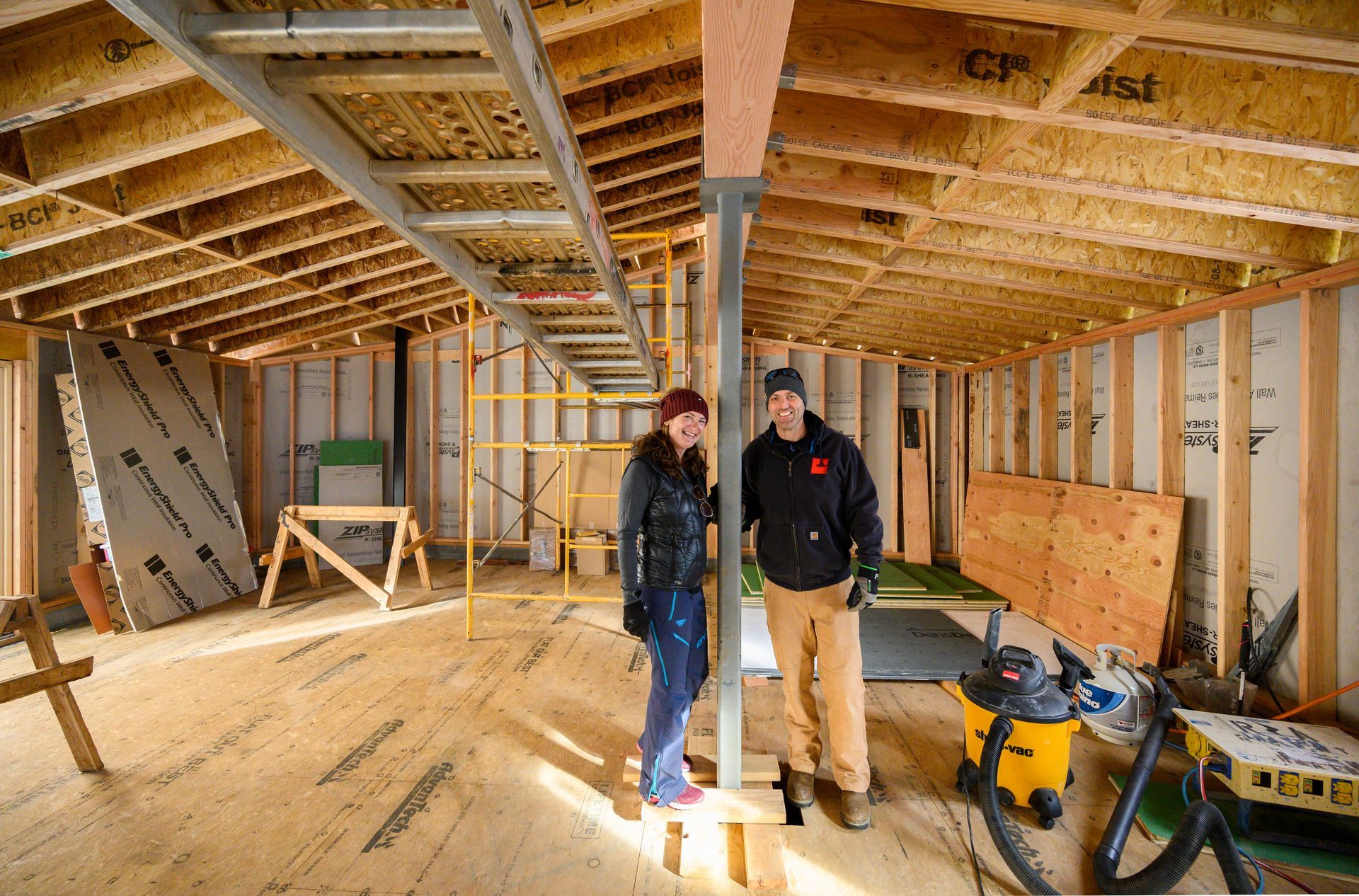 A woman in snowpants and a red beanie stands next to a man in a black sweatshirt inside a home under construction.