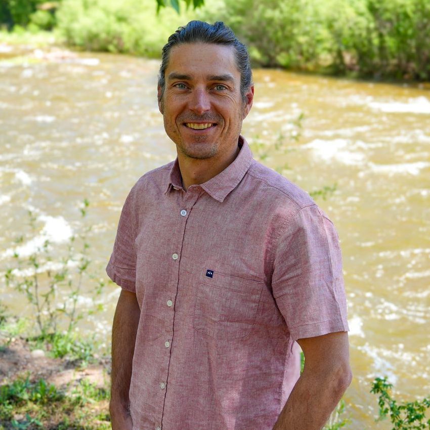 Lee has his hair pulled back, is wearing a red shirt, and is standing in front of a river. 