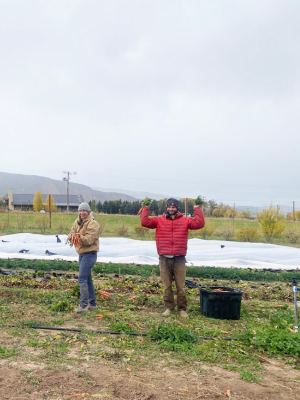 Two gardener's hold up their harvest at a sustainable farm with mountains in the background.