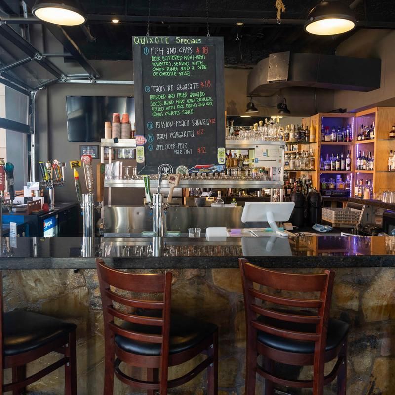 Empty bar interior with wooden stools, a chalkboard menu, and shelves of bottles behind the counter