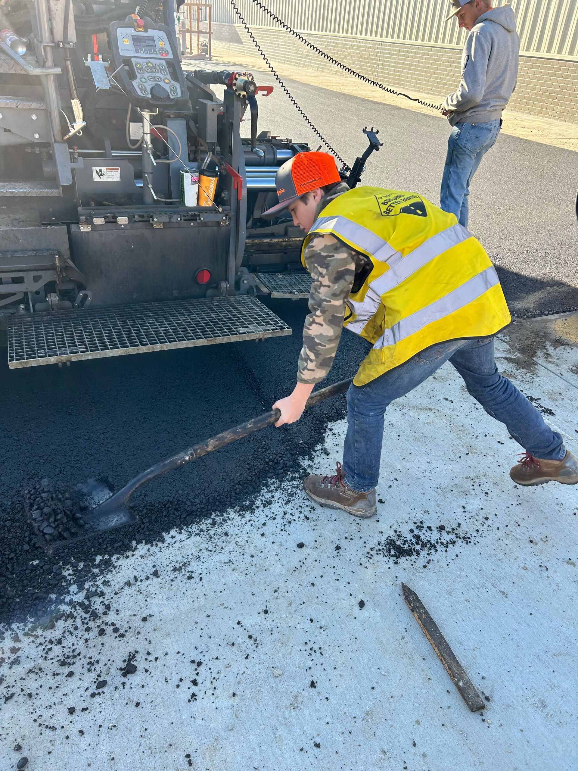 A man wearing a yellow vest and orange hat is working on a road