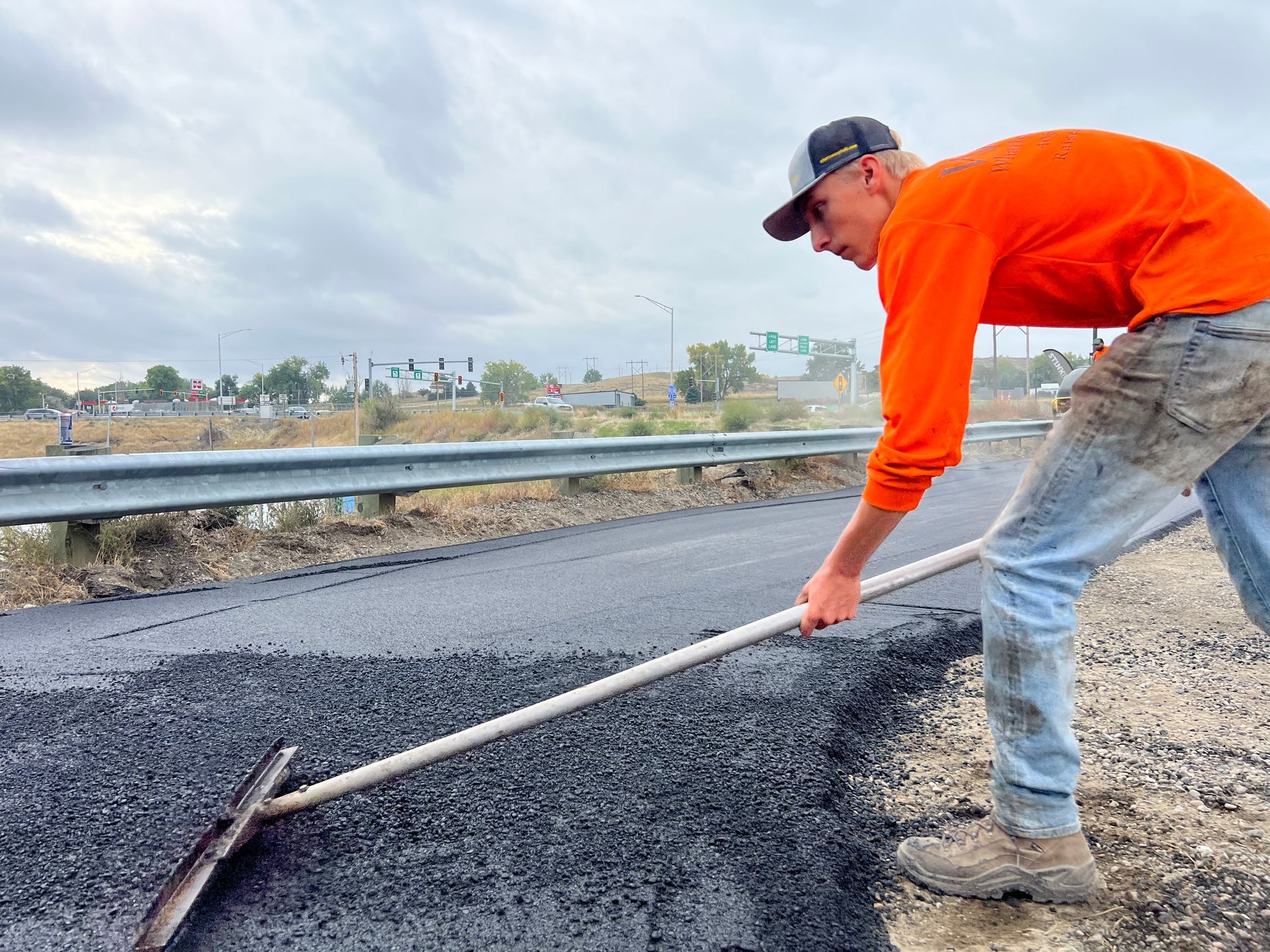 A man in an orange shirt is raking a road with a rake.