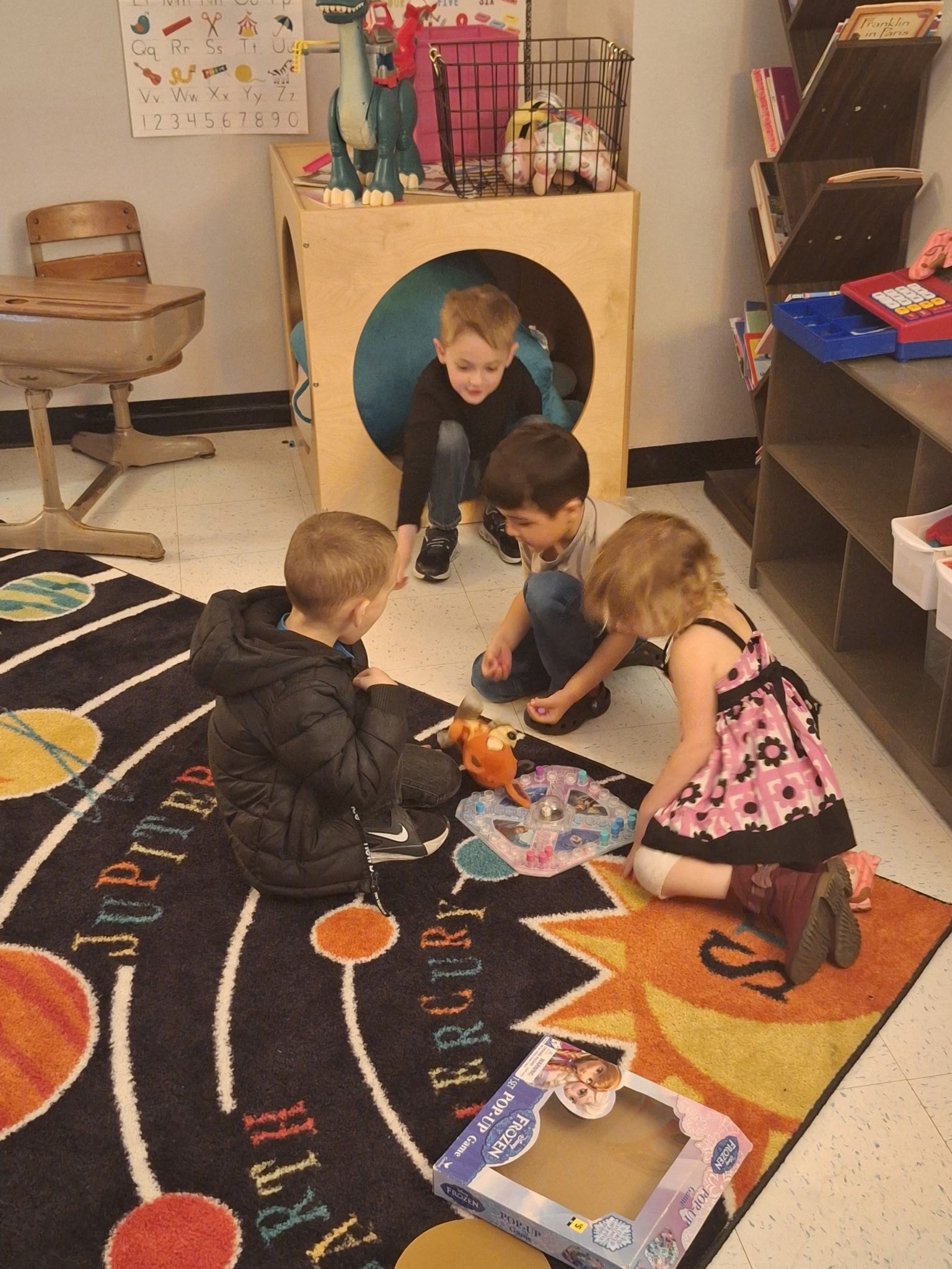 Four children playing with a small toy on a rug featuring a solar system design inside a classroom.