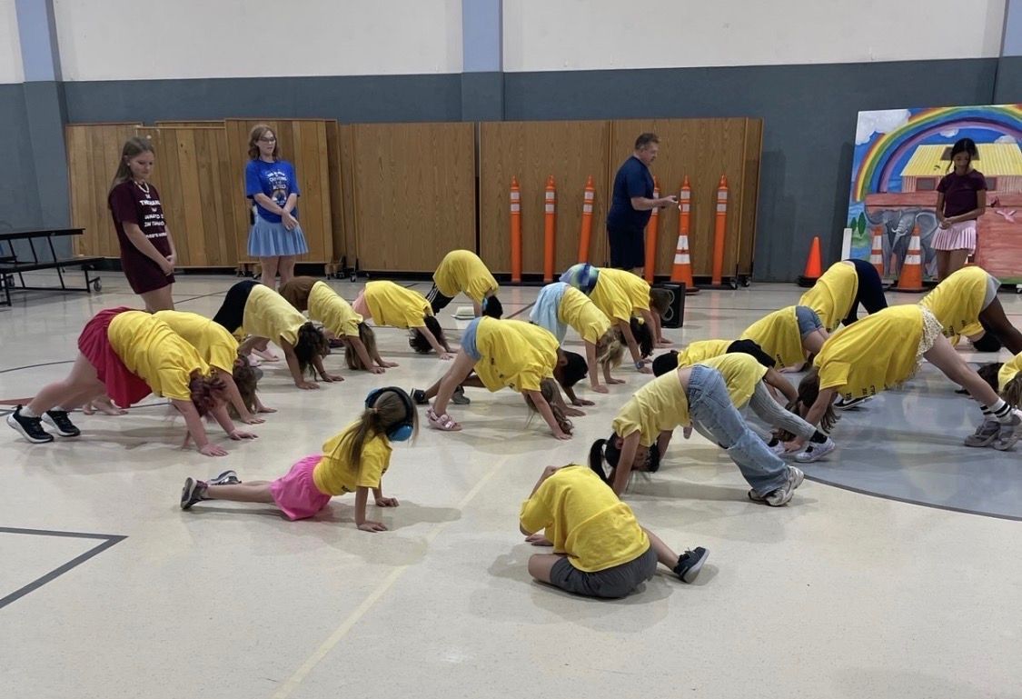 A group of children in yellow shirts performs yoga poses in a large indoor activity room with adults supervising nearby.