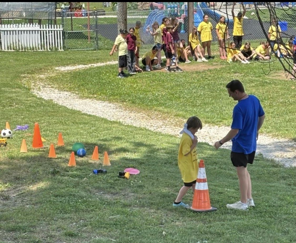 An adult in a blue shirt guides a child in a yellow shirt at an outdoor sports camp with cones and balls on the grass.