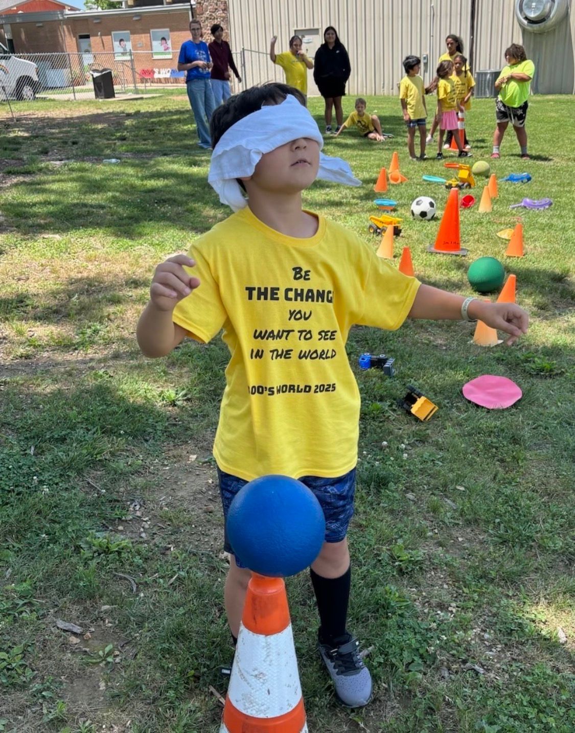 A blindfolded child in a yellow shirt with a blue ball on a cone in an outdoor field, surrounded by children and cones.