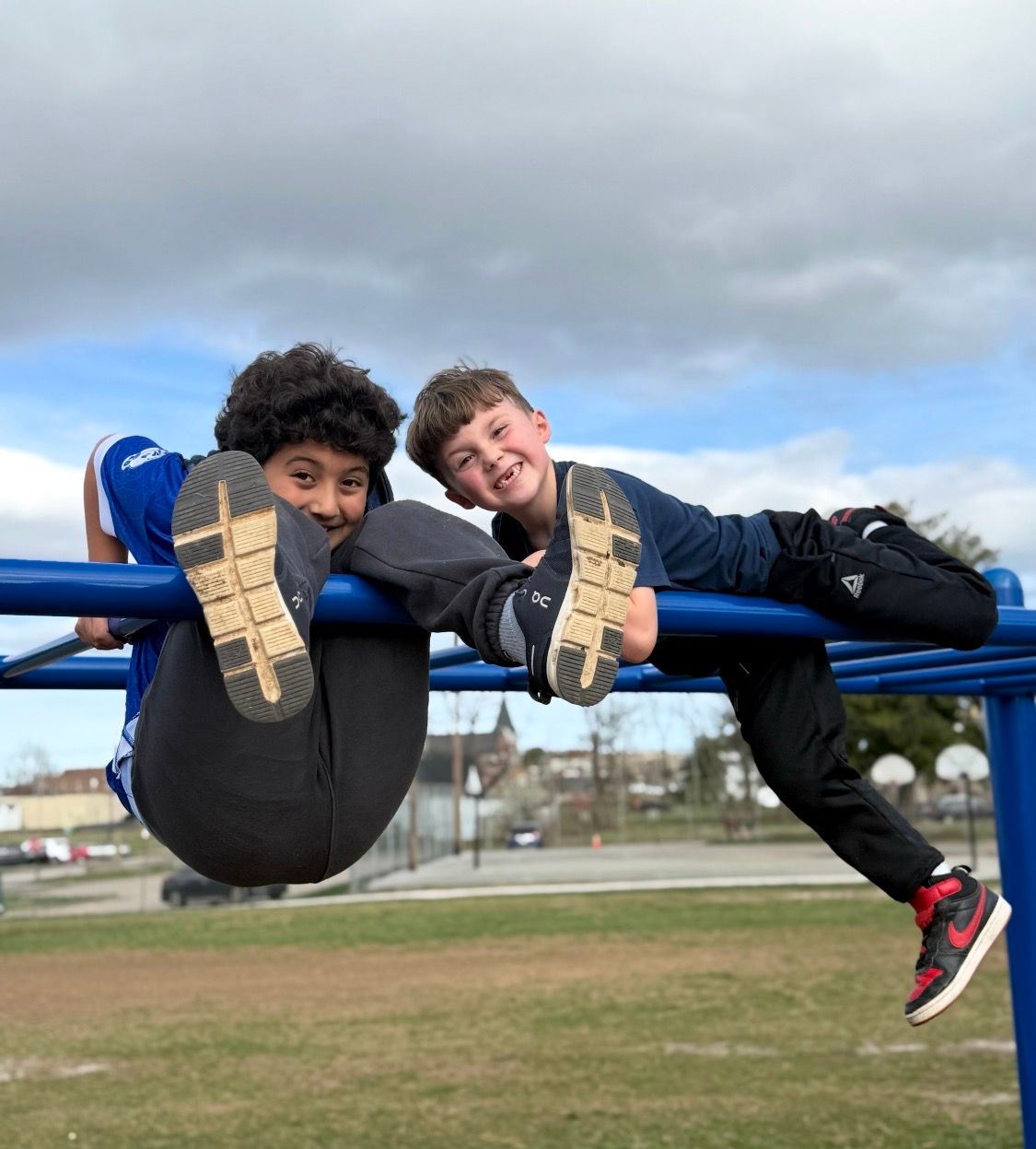 Two children hanging from blue monkey bars on a playground, smiling at the camera against a cloudy sky.