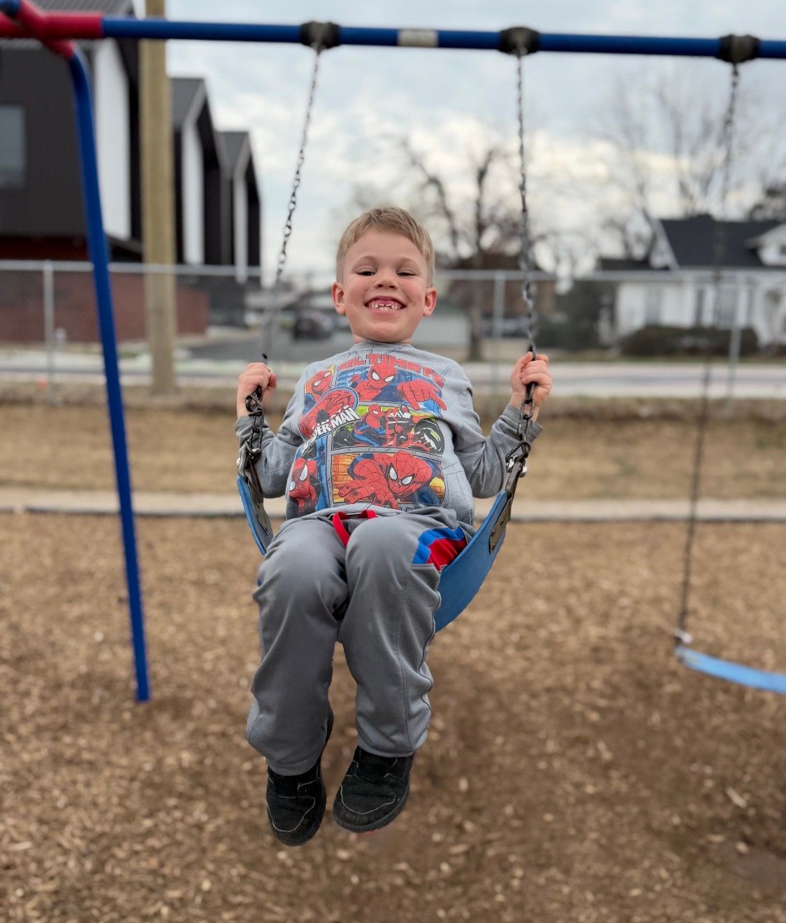 A smiling child wearing a Spider-Man shirt sits on a blue playground swing at an outdoor park.