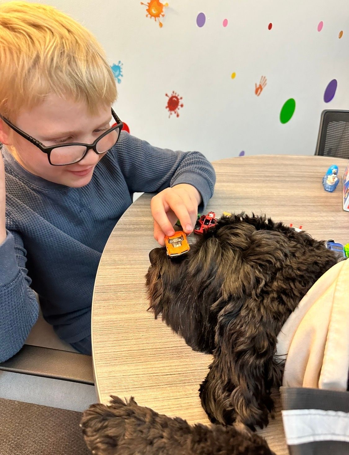A person with glasses plays with a small orange toy car on the nose of a resting black dog at a table.