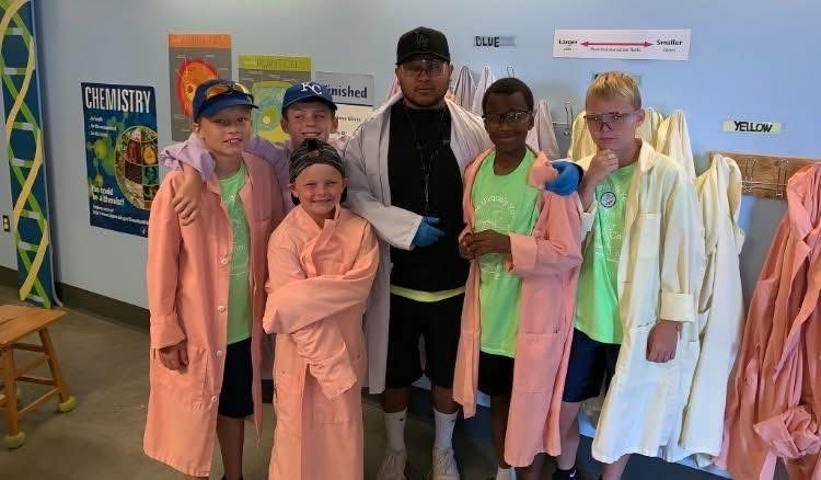 A group of five children and one adult standing in a room, wearing lab coats and bright green shirts.