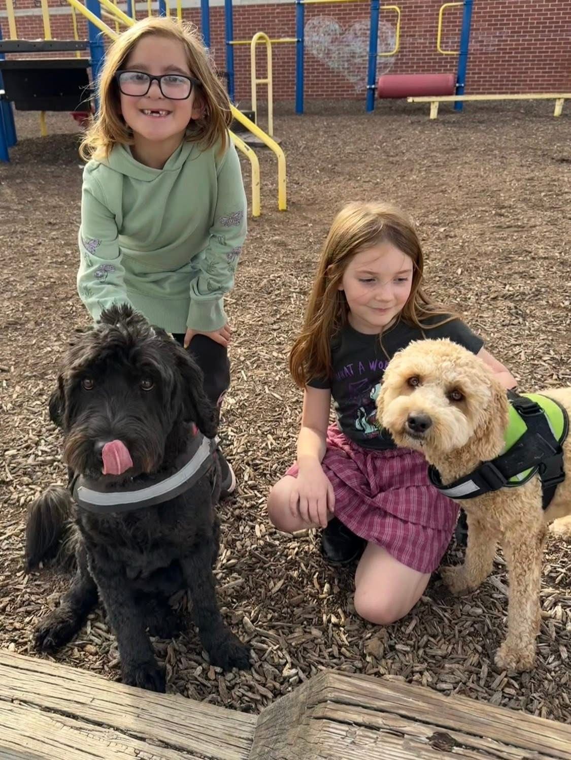 Two children pose with a black dog and a tan dog on a playground, with the black dog licking its nose.