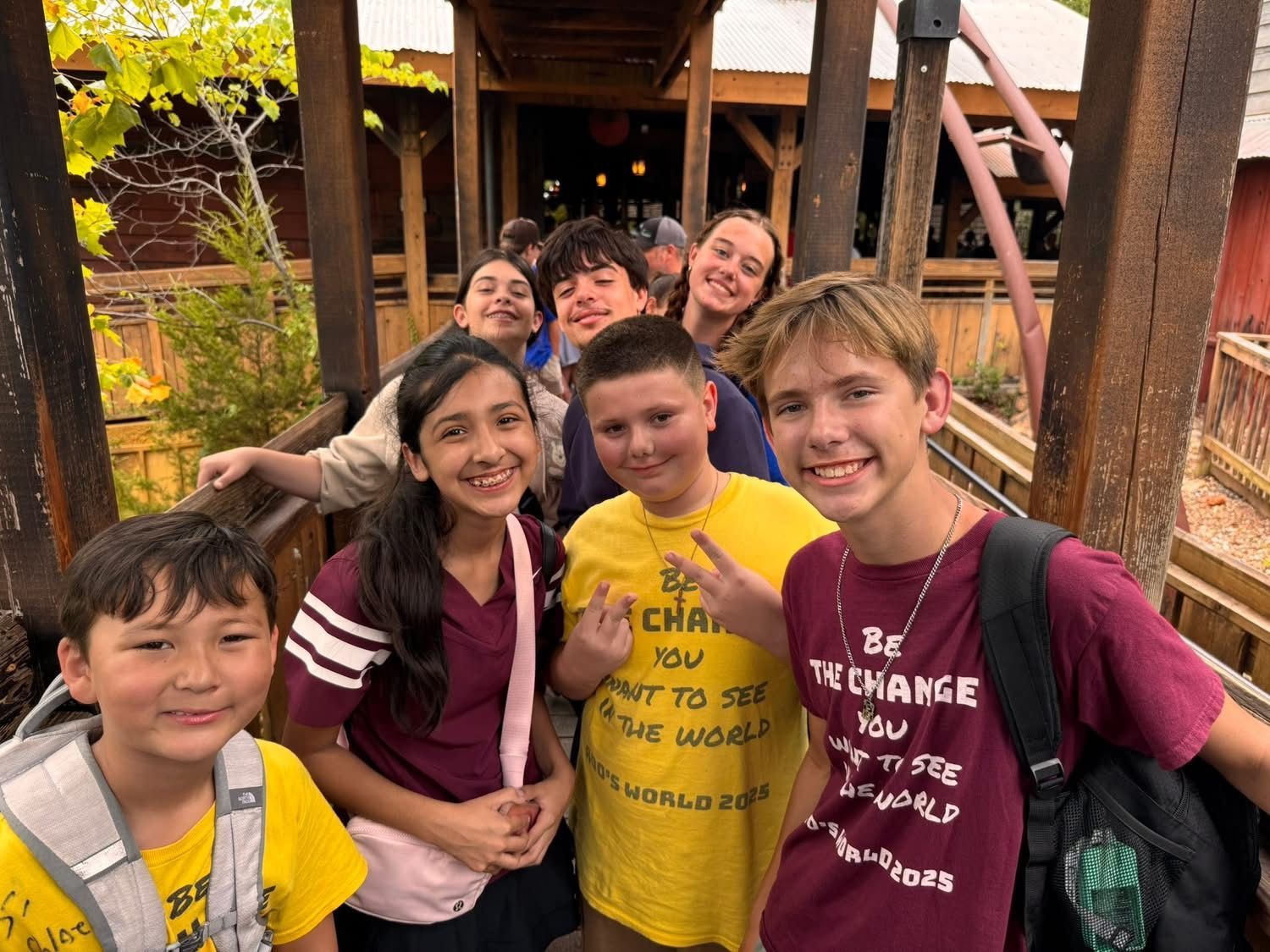 A group of students smile in an outdoor wooden walkway, some wearing yellow or maroon shirts, with a roller coaster nearby.