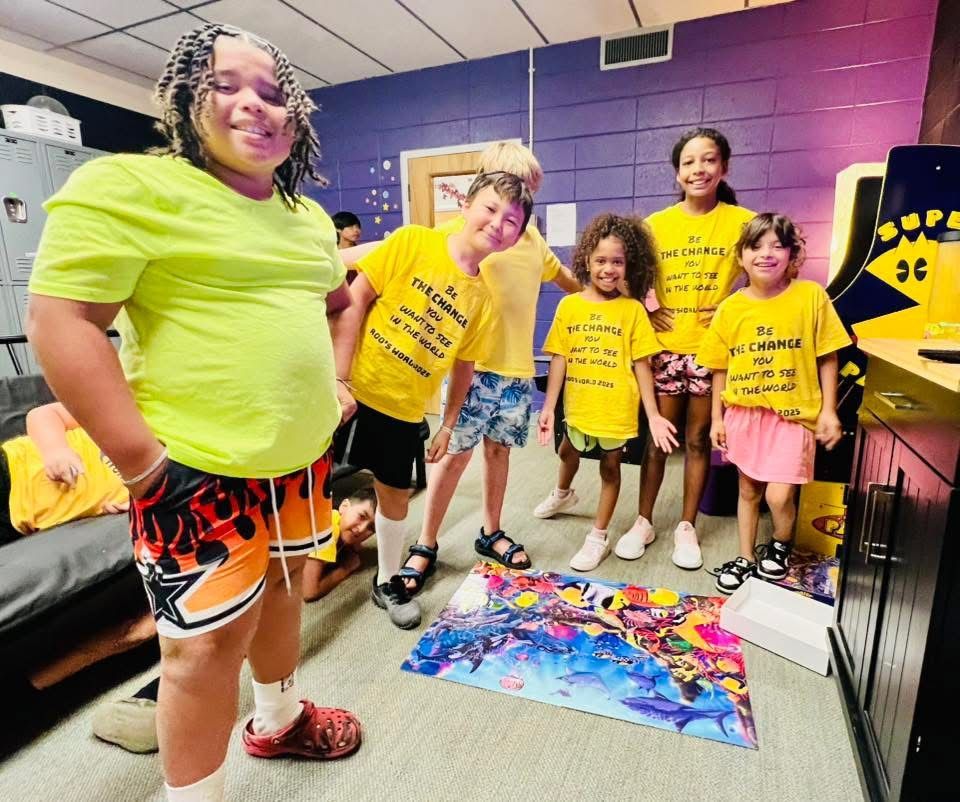Group of people in masks, some wearing yellow shirts, smiling at the camera in a brightly lit room.