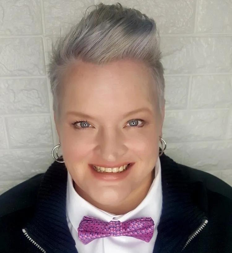 Woman with short gray hair, smiling, wearing a white shirt, pink bow tie, black jacket, and hoop earrings.