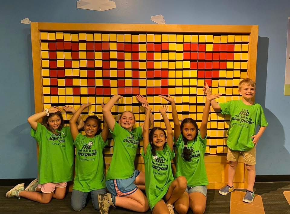 Six children wearing green shirts pose in front of a wall of yellow and red blocks that spell 