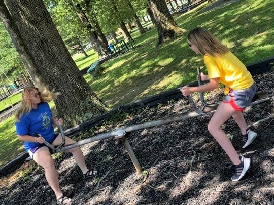 Two people in a park playing on a seesaw, one wearing a blue shirt and the other a yellow shirt.