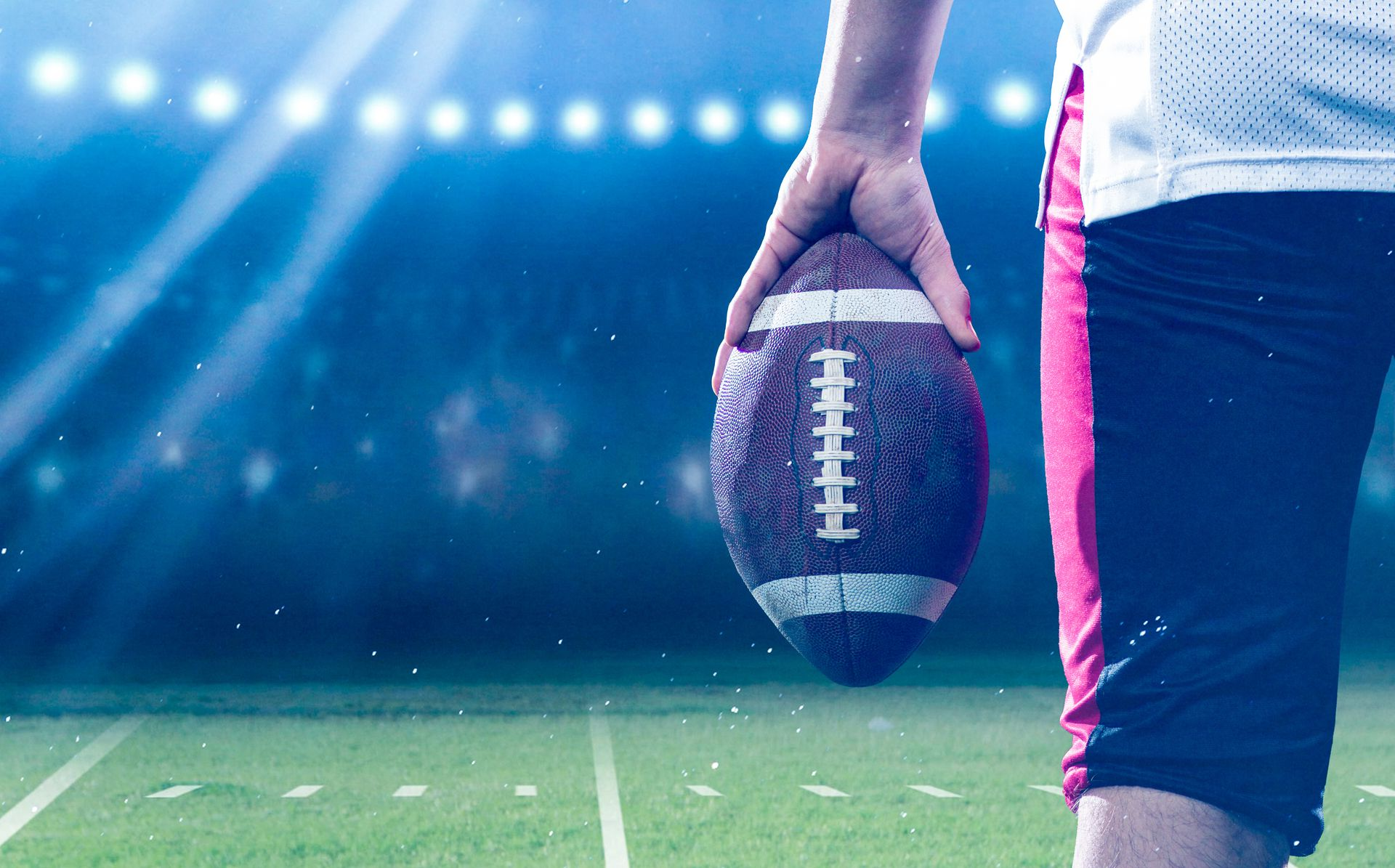 Football player holding a football on a green field at a stadium at night.