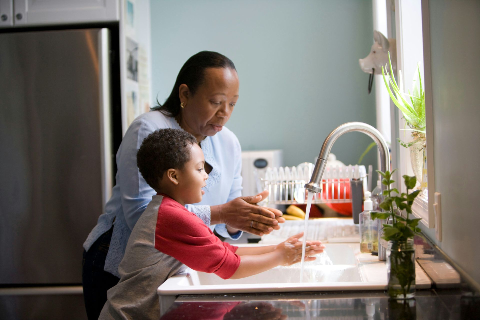Woman and young boy wash hands together at a kitchen sink.