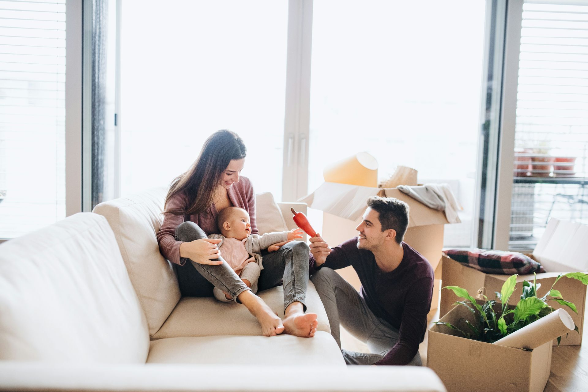 Family on a couch in new home with moving boxes: mother with baby, father interacting. Bright, sunny room.