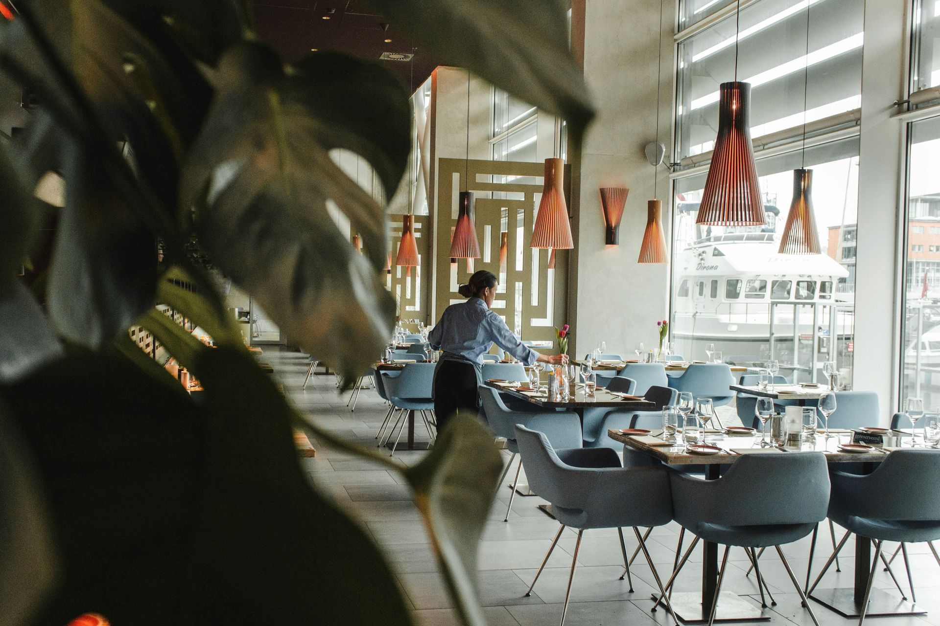 Restaurant interior, server setting tables. Blue chairs, large windows, orange pendant lights, tropical foliage.
