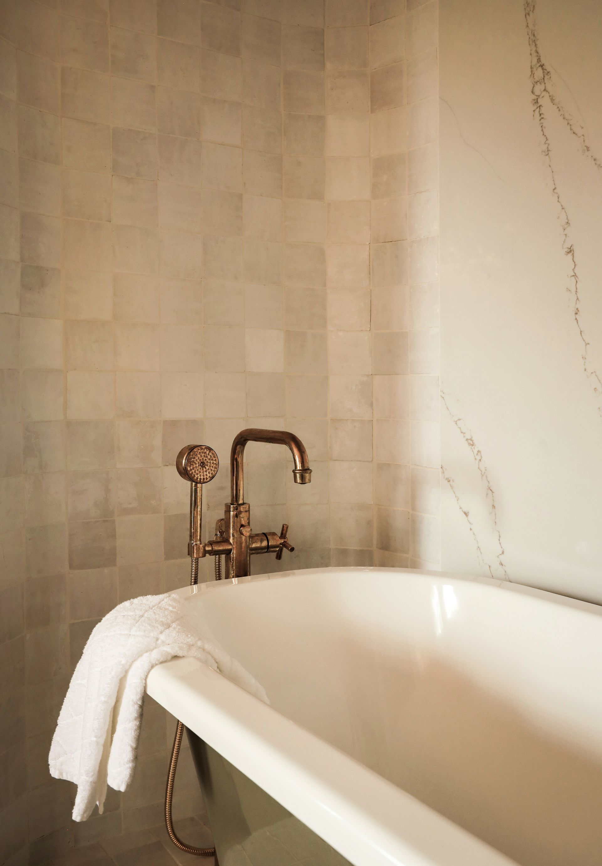 Copper faucet and white bathtub in a bathroom, white towel, light gray tiled walls.